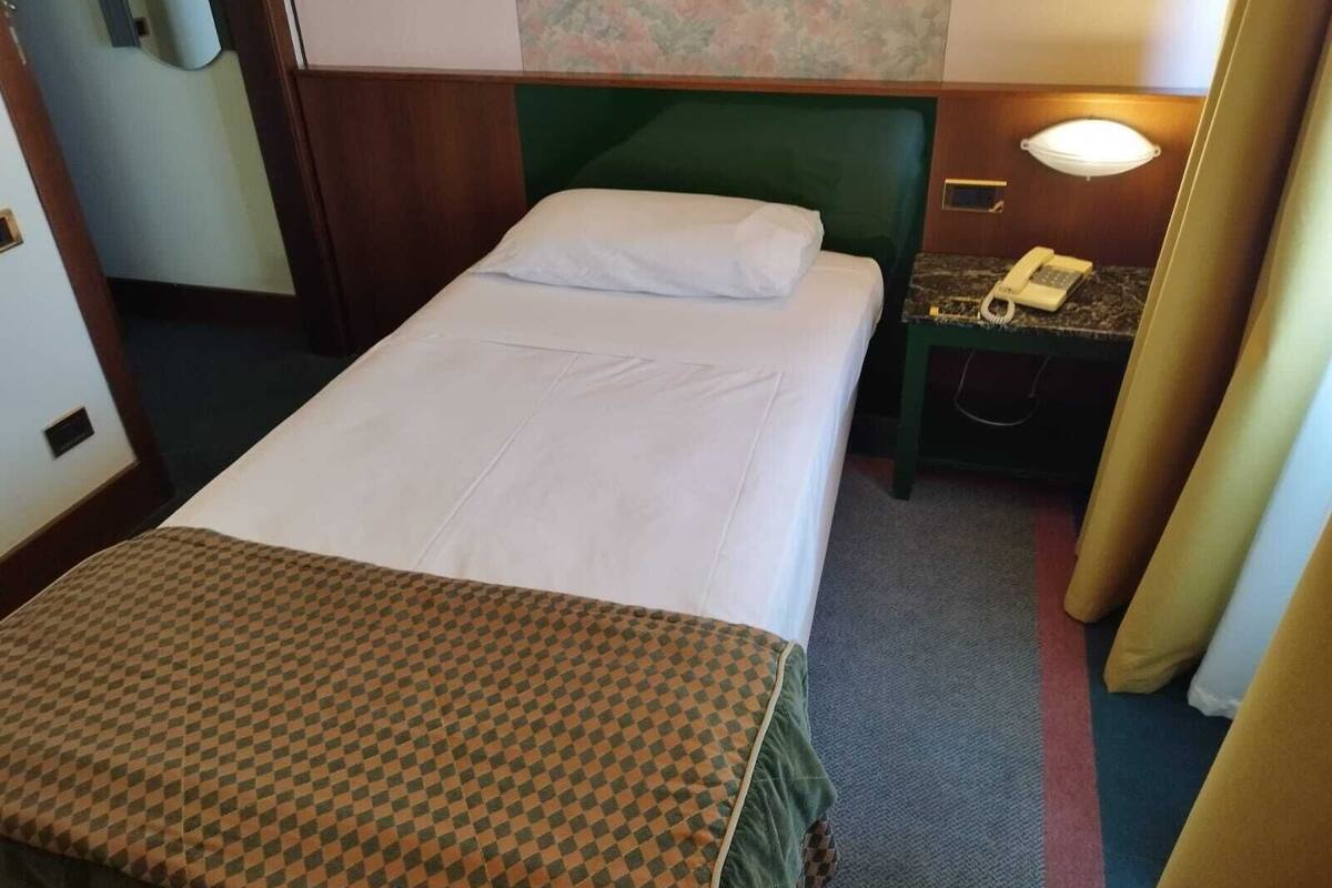 A hotel room with a single bed, a nightstand with a telephone, and a wall-mounted lamp.