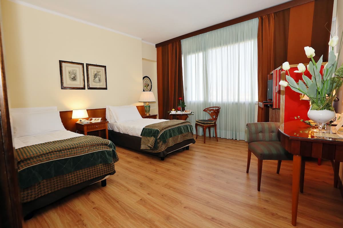 Une chambre d’hôtel avec deux lits, un bureau avec une chaise, une petite table avec une lampe et une grande fenêtre avec des rideaux.