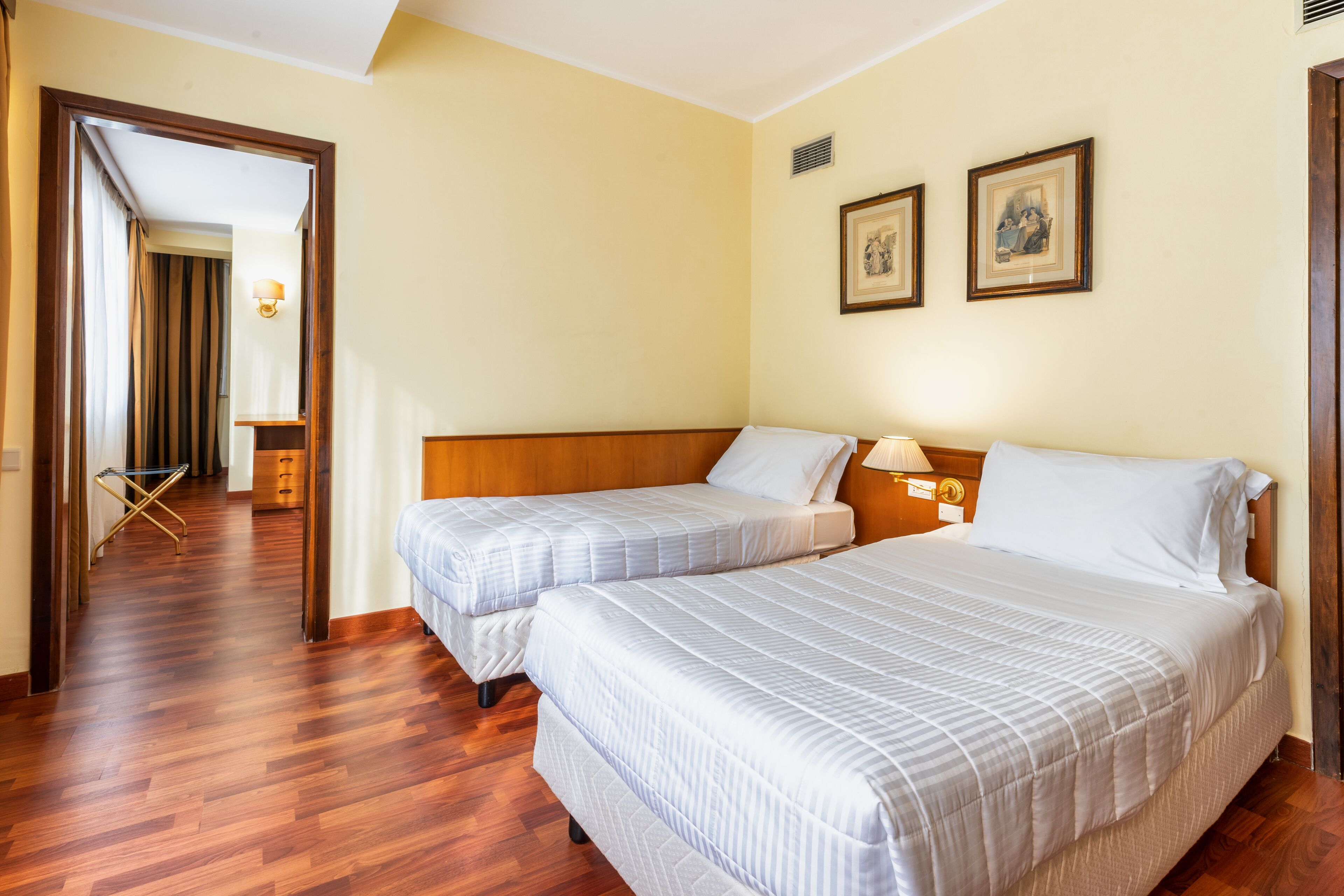 Une chambre d’hôtel avec deux lits, un plancher en bois, une tête de lit en bois, une table de chevet avec une lampe et une porte donnant sur un couloir.