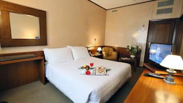 Habitación de hotel con cama, escritorio, silla, televisor y mesita de noche con lámpara.