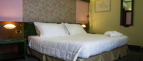 A neatly made bed with a white comforter and pillows, a green nightstand with a lamp, a floral headboard, and a wall-mounted mirror.