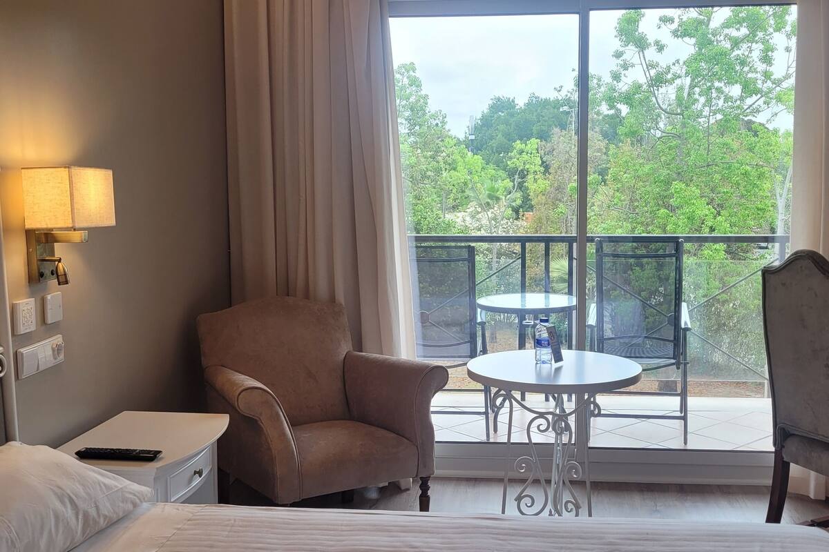 Habitación de hotel con cama, una silla, escritorio, una mesita y balcón con vista a árboles.