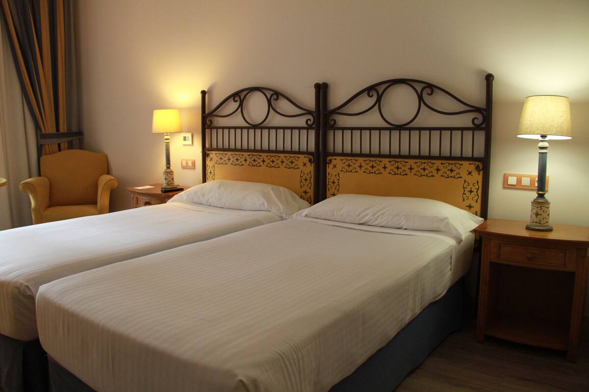 Habitación de hotel con dos camas individuales, una silla, dos mesitas de noche con lámparas y una cortina.