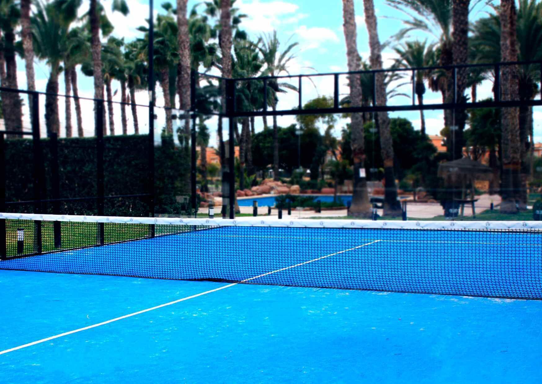 A tennis court with a blue surface and net, surrounded by palm trees and a pool in the background.