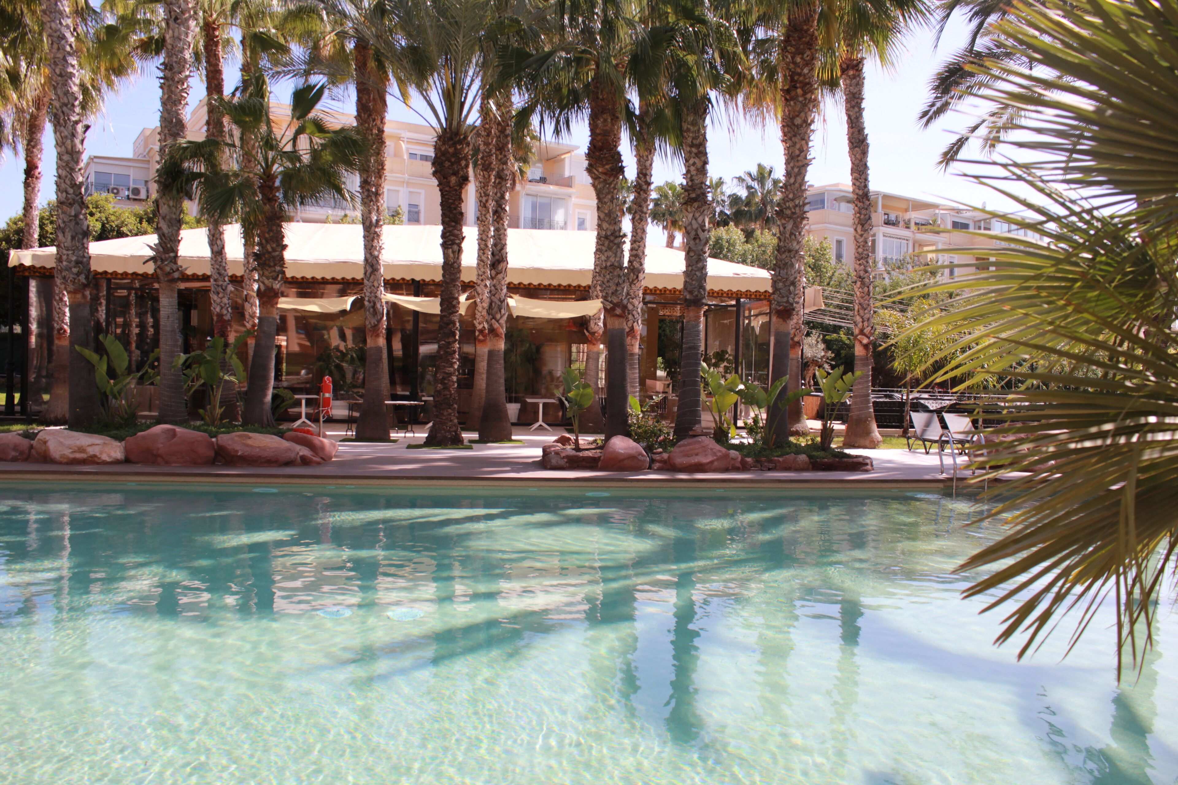 A pool area with palm trees, a building with a beige roof, and a clear blue pool.