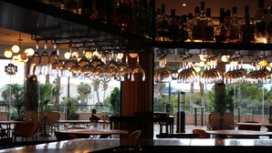 A bar area with hanging wine glasses and bottles, outdoor seating with tables and chairs, and a view of palm trees and buildings. - Hotel Alicante Golf (Alicante)