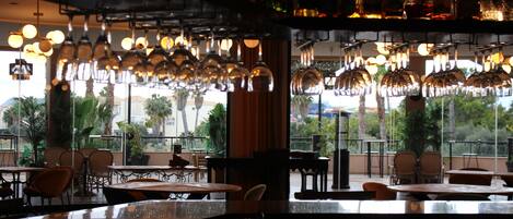 Zona de bar con copas y botellas de vino colgando, mesas y sillas para exterior, y vistas a palmeras y edificios.