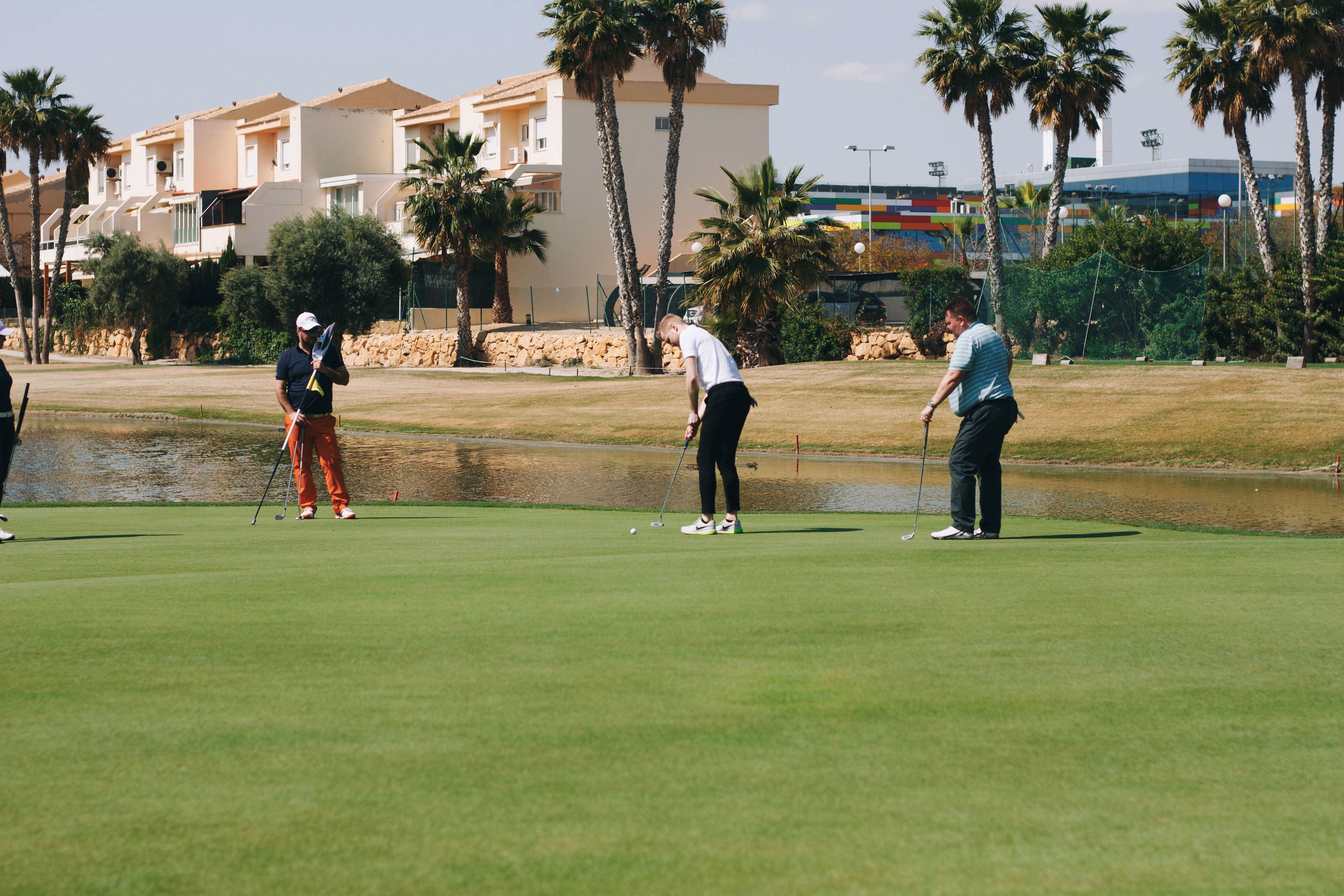 A golf course with a water hazard, palm trees, and a building in the background.