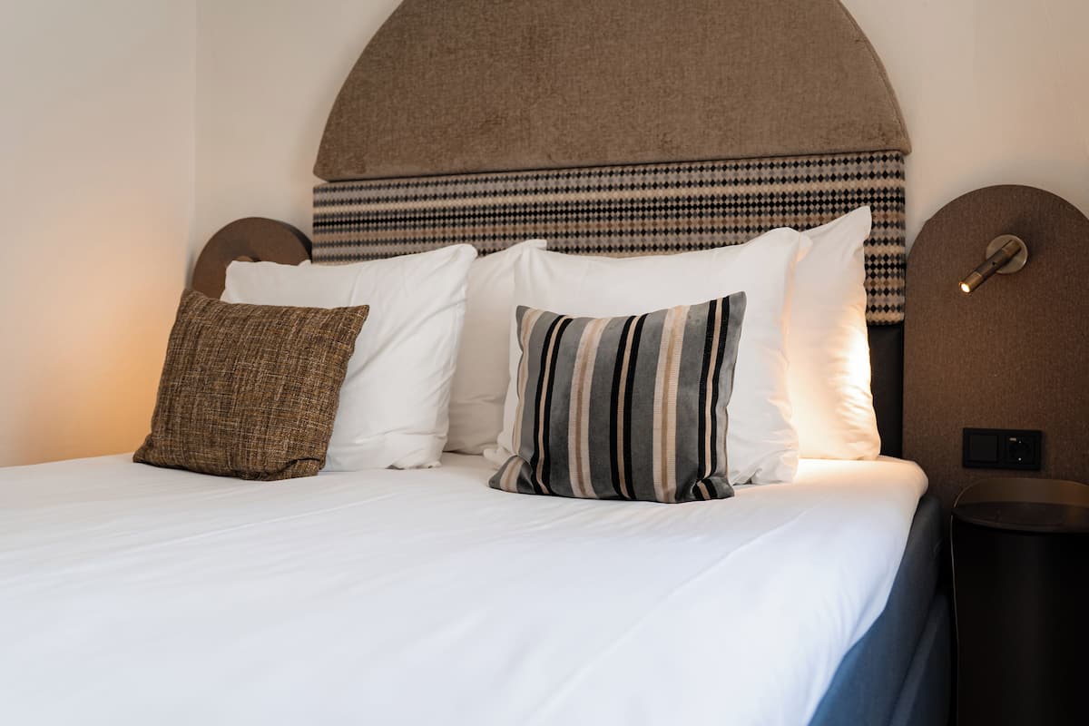Un lit bien fait, avec des draps blancs et des oreillers à rayures, une tête de lit à motif pied-de-poule et une petite table de chevet noire avec une lampe.