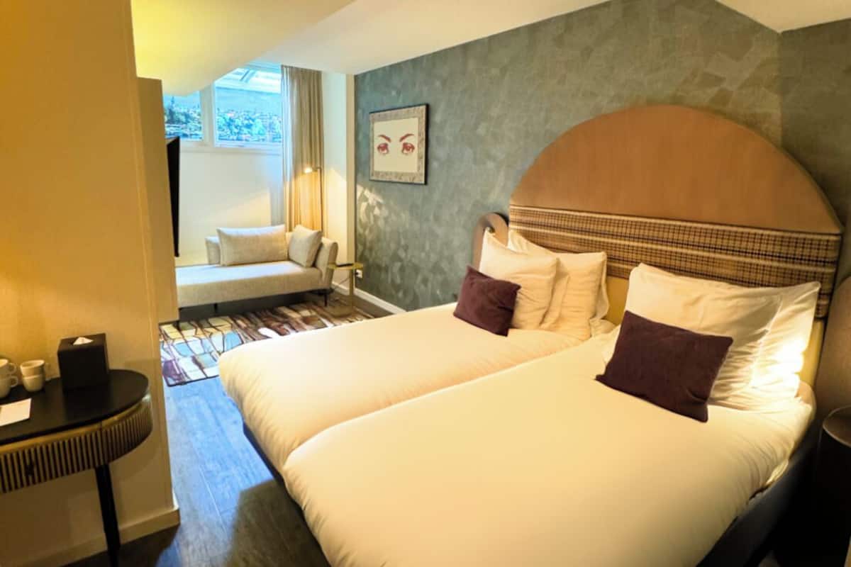 Un lit double avec une tête de lit en bois, deux oreillers, une table de chevet avec une lampe et une fenêtre donnant sur l’extérieur.