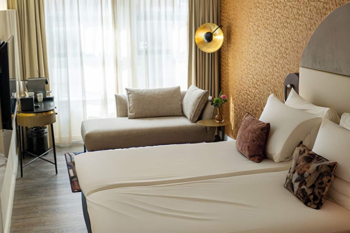 Une chambre d’hôtel avec un grand lit, une table de chevet avec une lampe, une petite table avec une machine à café et une vue sur la ville par la fenêtre.