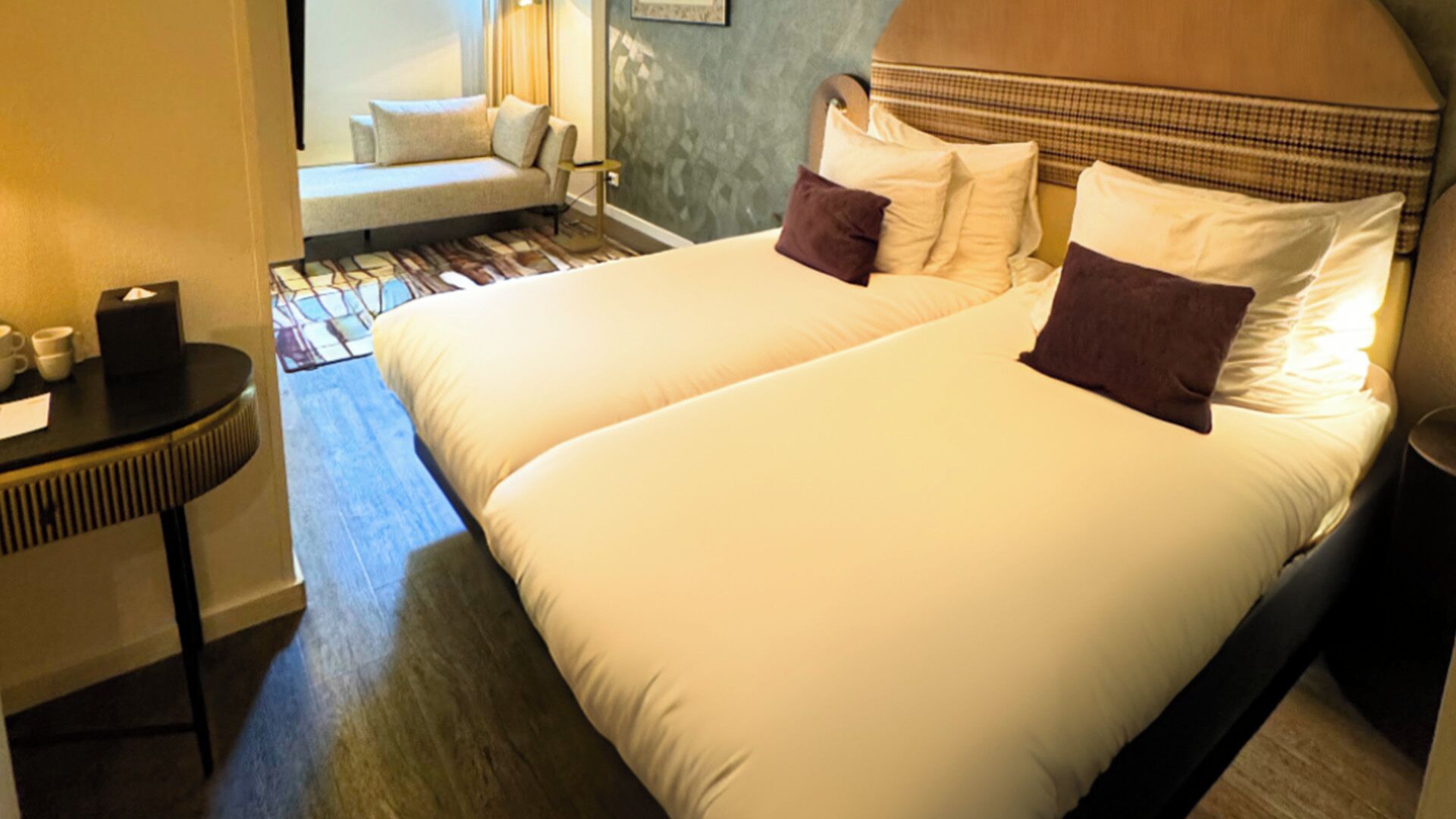 A hotel room with two beds, a small table with a speaker, and a sofa in the background.