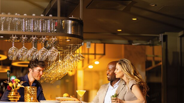 Een stel zit aan de bar, met een decoratieve kroonluchter, een menukaartstandaard en gestreept beklede barkrukken.
