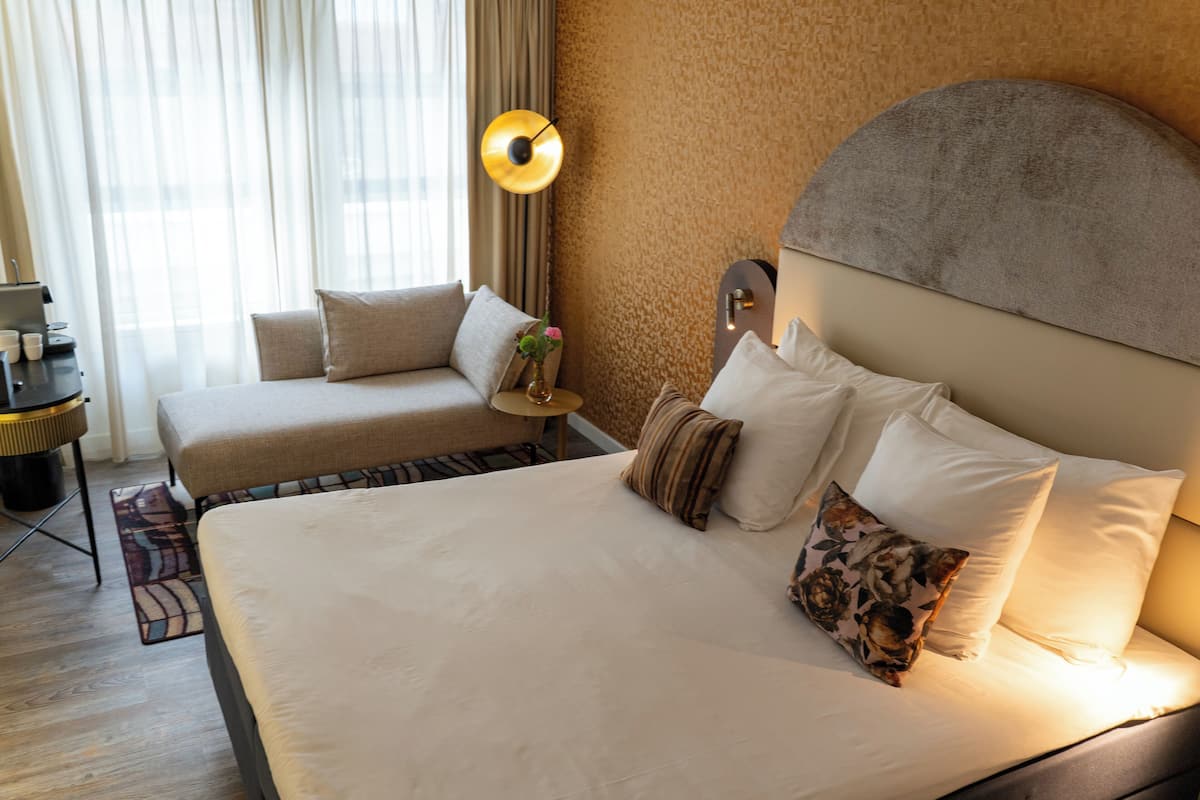 Une chambre d’hôtel avec un grand lit, un canapé, une petite table d’appoint avec une lampe, et une vue sur l’extérieur à travers des rideaux légers.