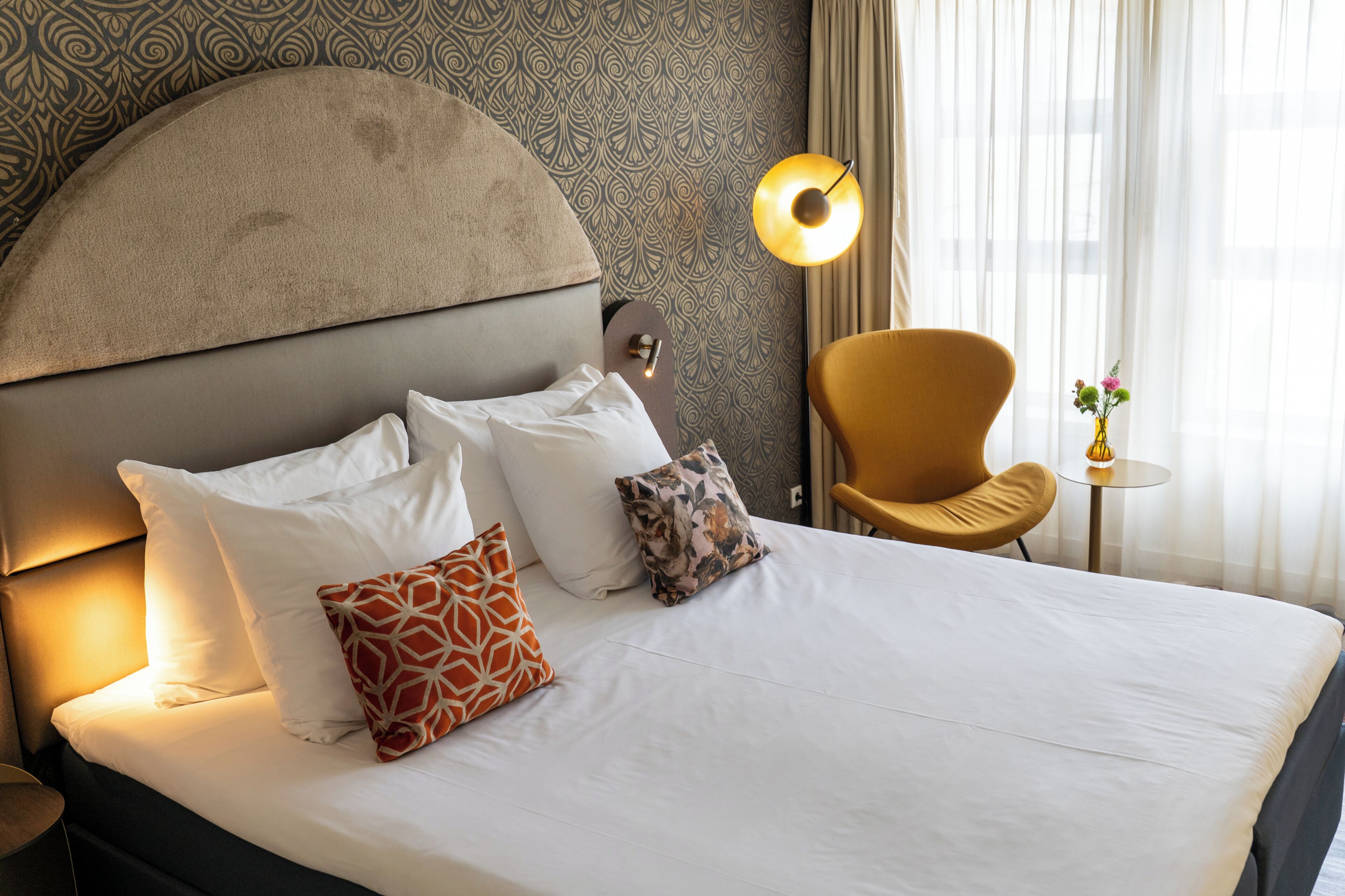 A hotel room with a large bed, a round table with a vase, a chair, and a patterned wall.