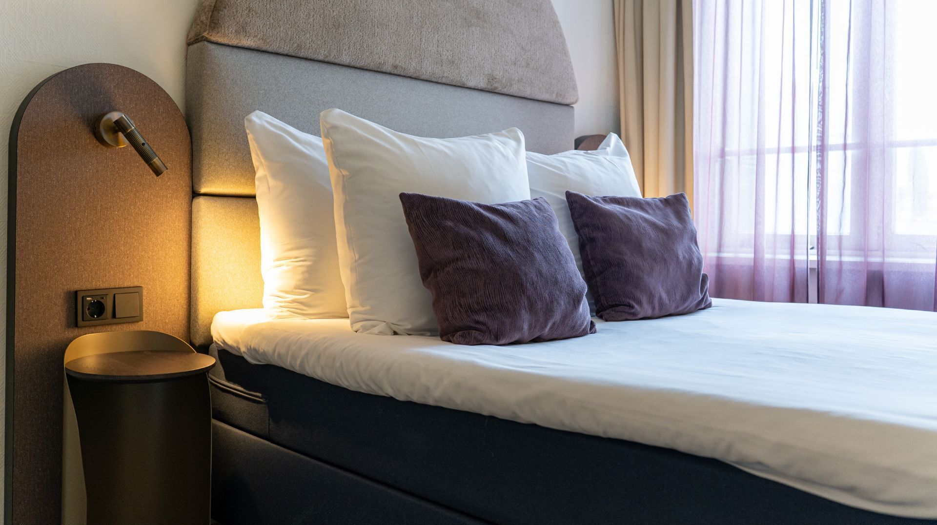 A hotel room with a bed, pillows, a headboard, and a bedside table.