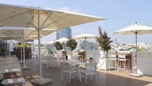 Una terraza en la azotea con mesas y sillas blancas, sombrillas grandes y vistas del horizonte urbano.