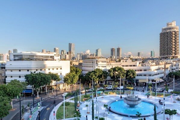Un paysage urbain avec des bâtiments modernes, une fontaine centrale et une zone piétonne agrémentée d’arbres et de bancs.