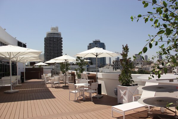 A rooftop terrace with white tables and chairs, umbrellas, and a view of the city skyline.