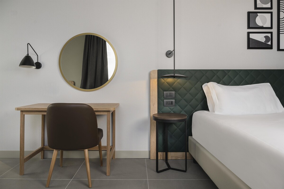 Una camera d'albergo con un letto, una scrivania, una sedia, uno specchio rotondo e una lampada a muro.