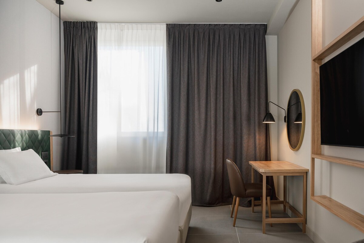 Una camera d'albergo con un letto, una scrivania, una sedia, una lampada e una televisione.