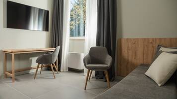 Una habitación moderna con un escritorio de madera, dos sillones grises, una cama con ropa de cama gris y un ventanal con cortinas.