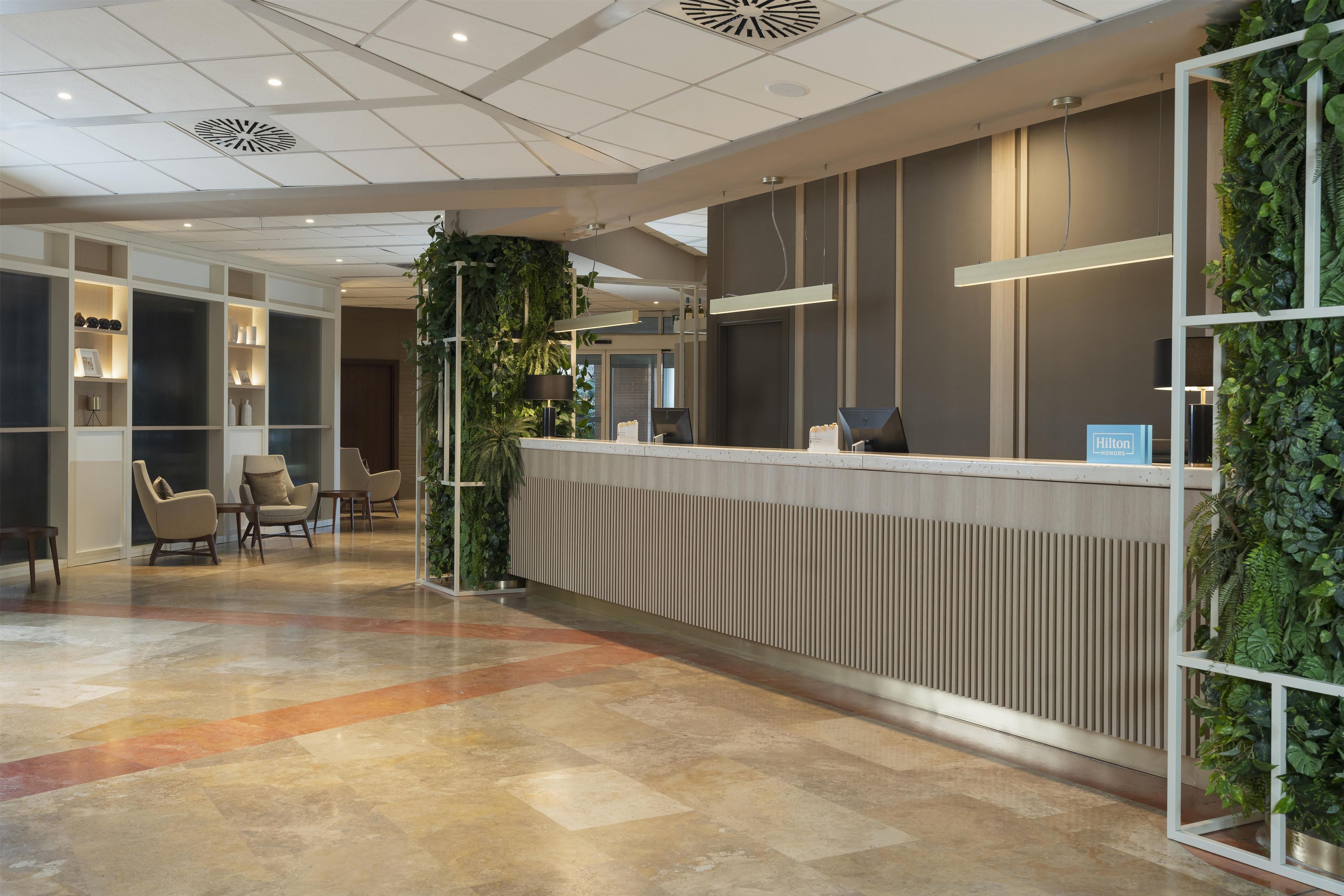Un'area di accoglienza hotel dal design moderno, con reception, zona salotto e piante ornamentali.