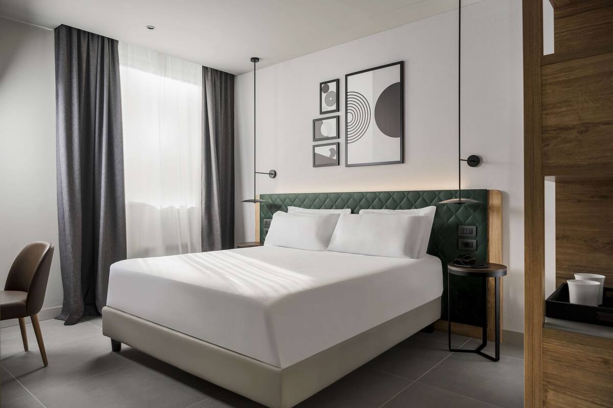 A modern hotel room with a large bed, a green upholstered headboard, a bedside table, and a chair.