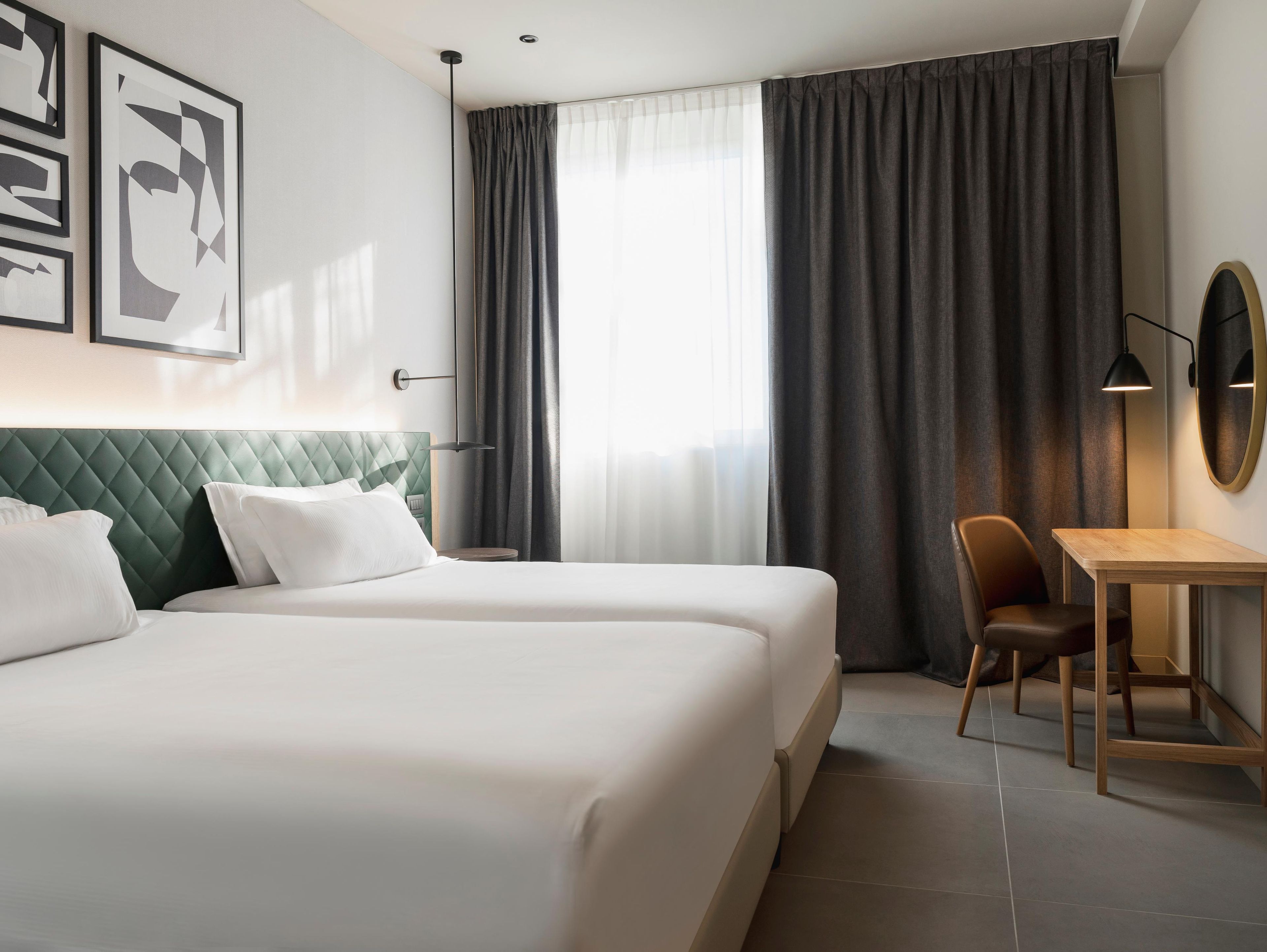 A hotel room with two beds, a desk, a chair, a large window with curtains, and framed artwork on the wall.