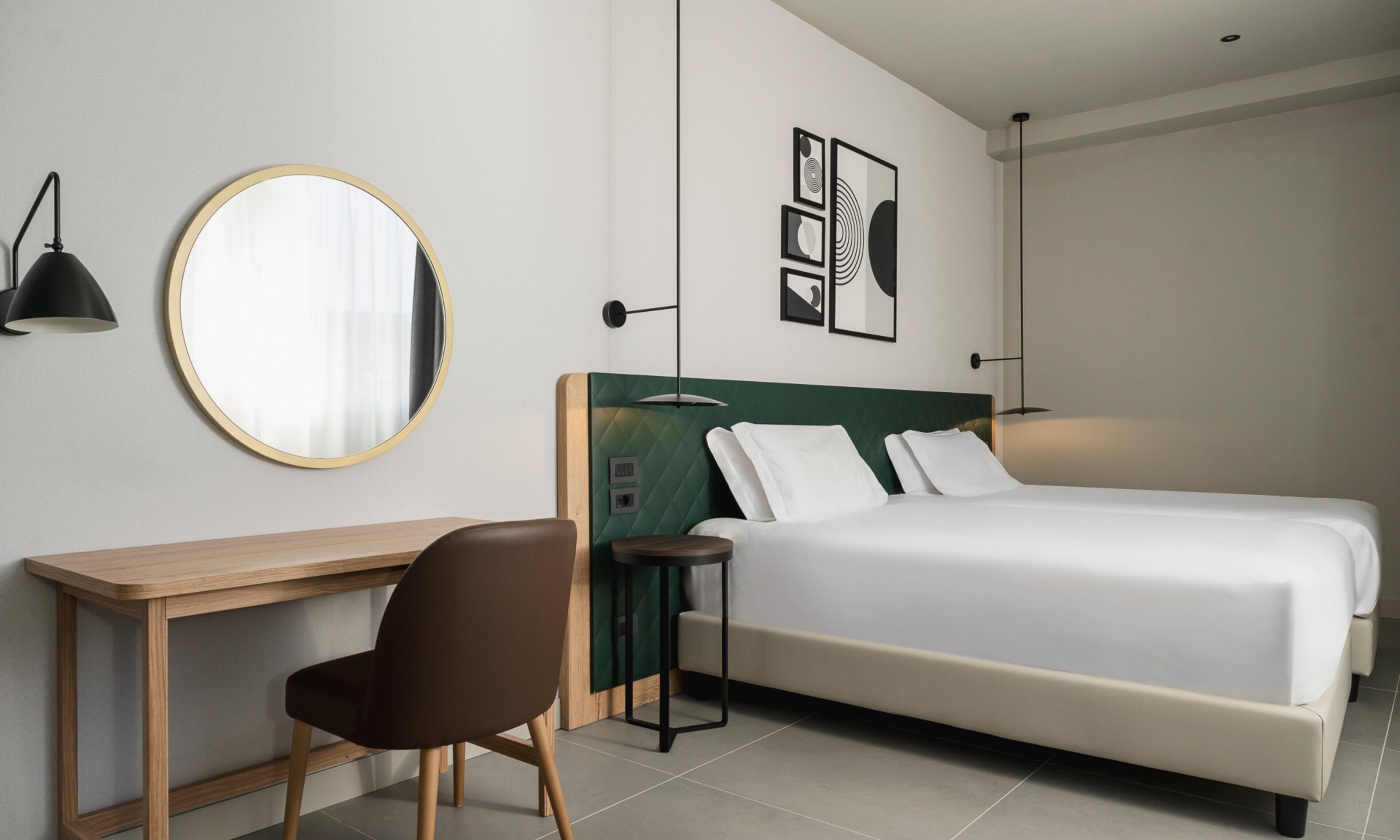 A hotel room with a bed, a desk, a chair, a round mirror, and wall art.