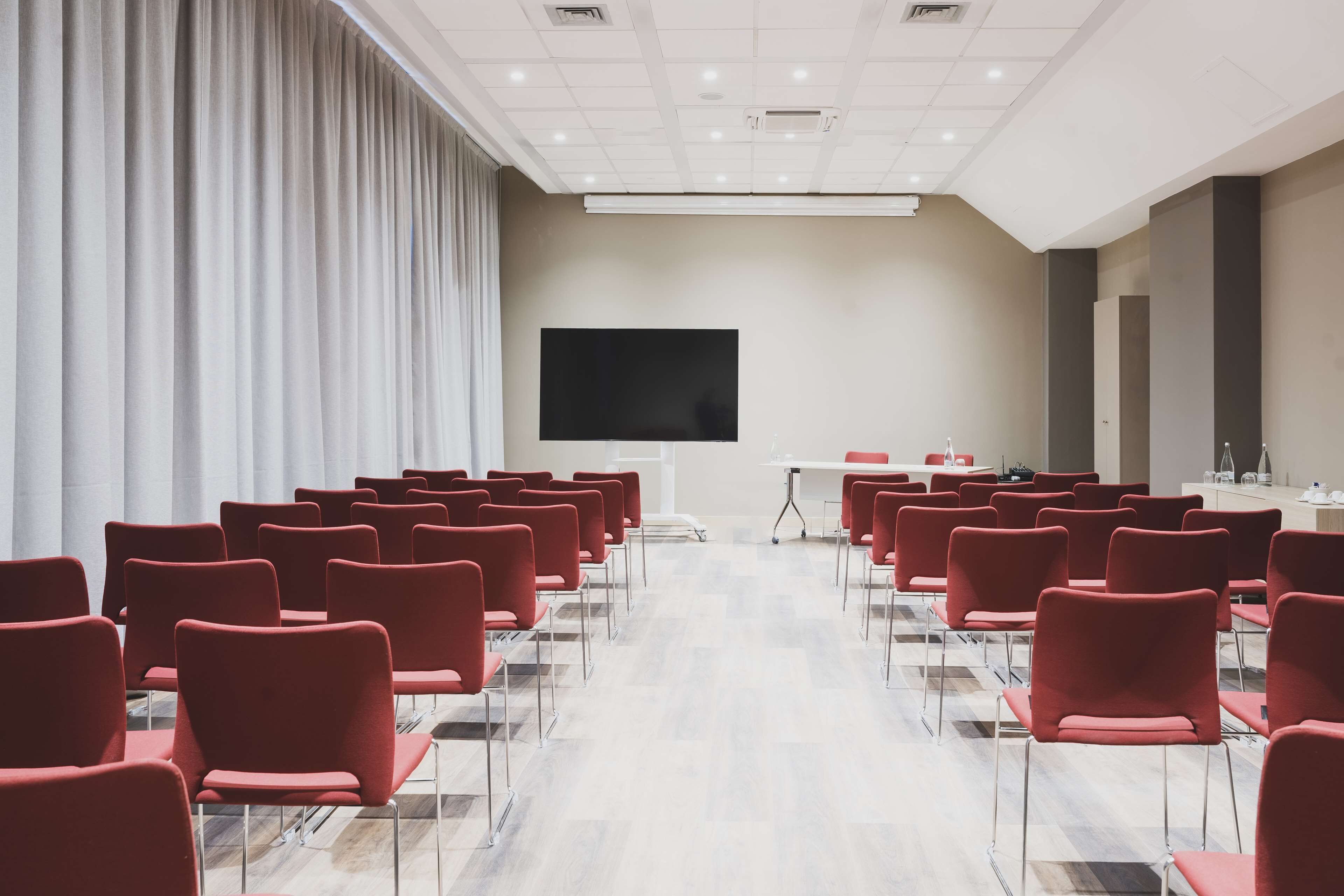 Een vergaderzaal met rijen rode stoelen, een grote flatscreen-tv en een witte tafel vooraan.