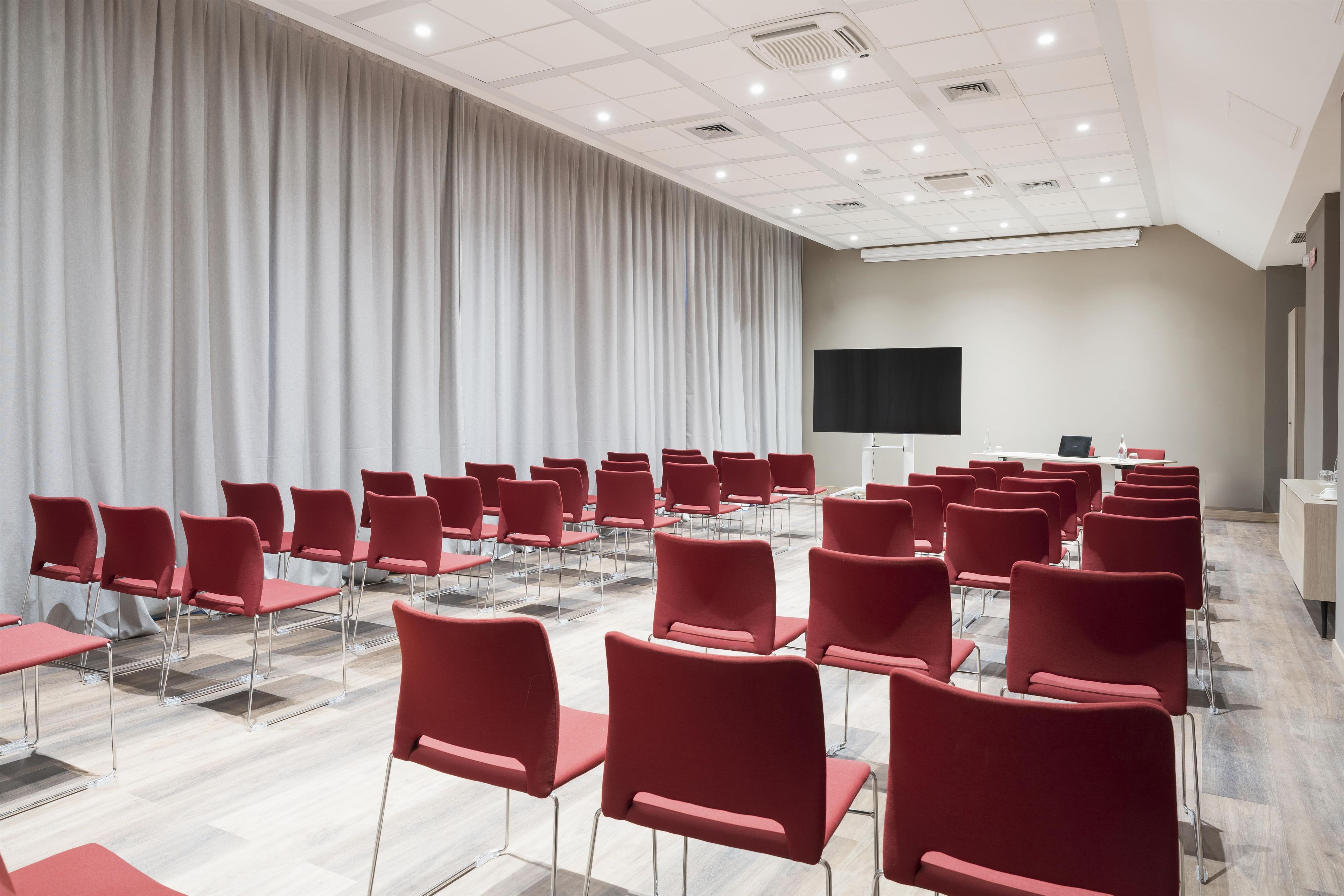 Ein Konferenzraum mit Reihen roter Stühle, einem großen weißen Vorhang, einem Flachbildfernseher und einer Decke mit Einbautenleuchten.