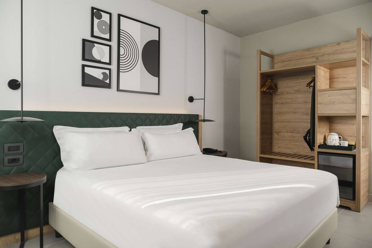 A neatly made bed with a green headboard, a wooden wardrobe with a mirror, and a wall with framed artwork.