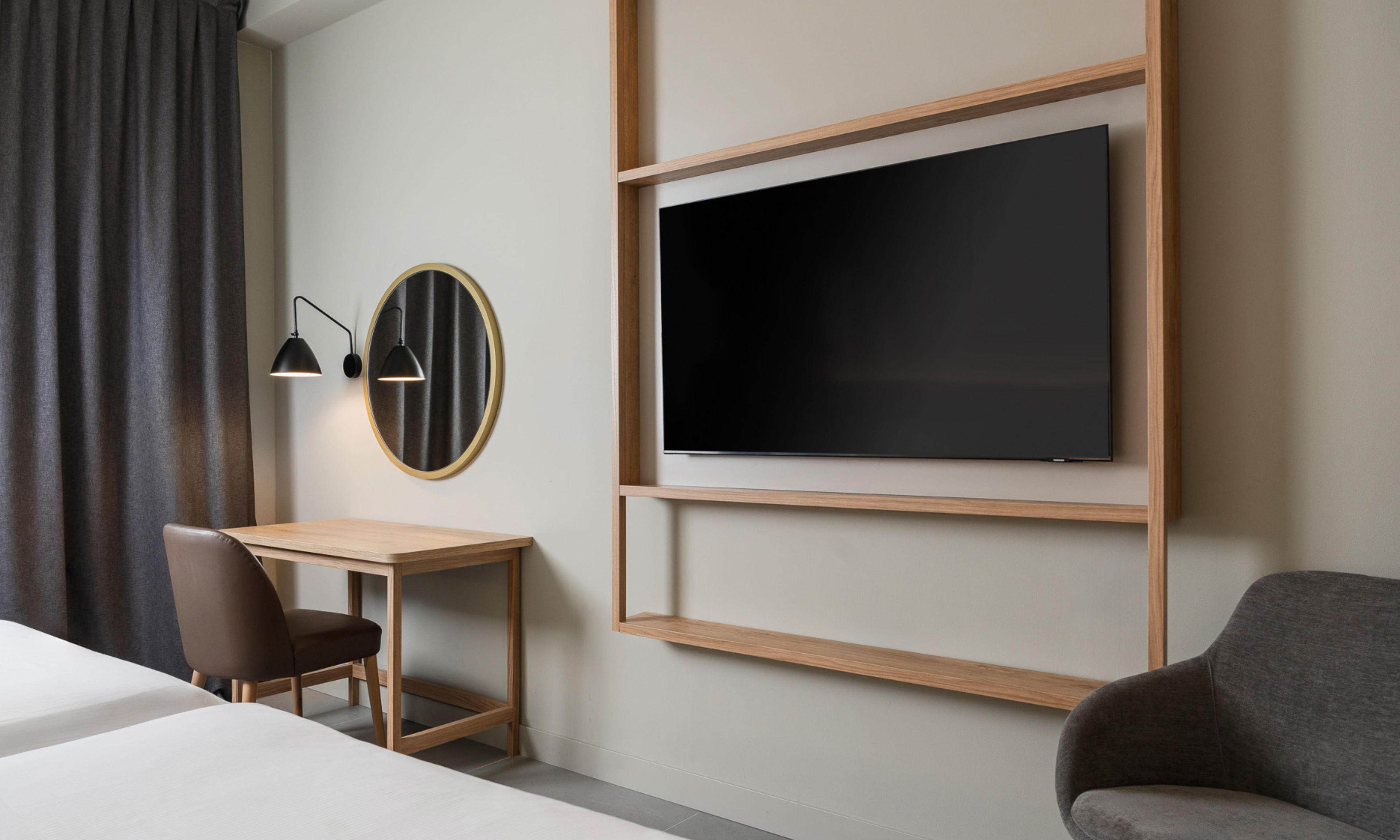 A hotel room with a bed, a desk, a chair, a TV mounted on the wall, and a window with curtains.