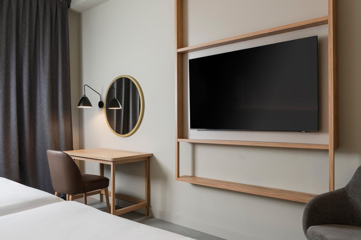 Camera d'albergo con un letto, una scrivania, una sedia, una TV a parete e una finestra con tende.