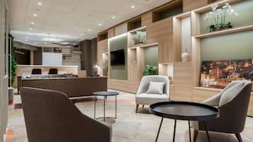 A modern lounge area with sofas, armchairs, and a small table, featuring a wooden wall with a television and decorative plants.