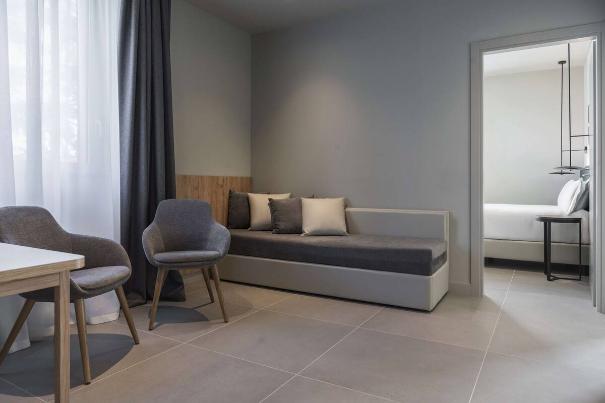 A modern, minimalist room with a sofa, two chairs, a small table, and a bed.