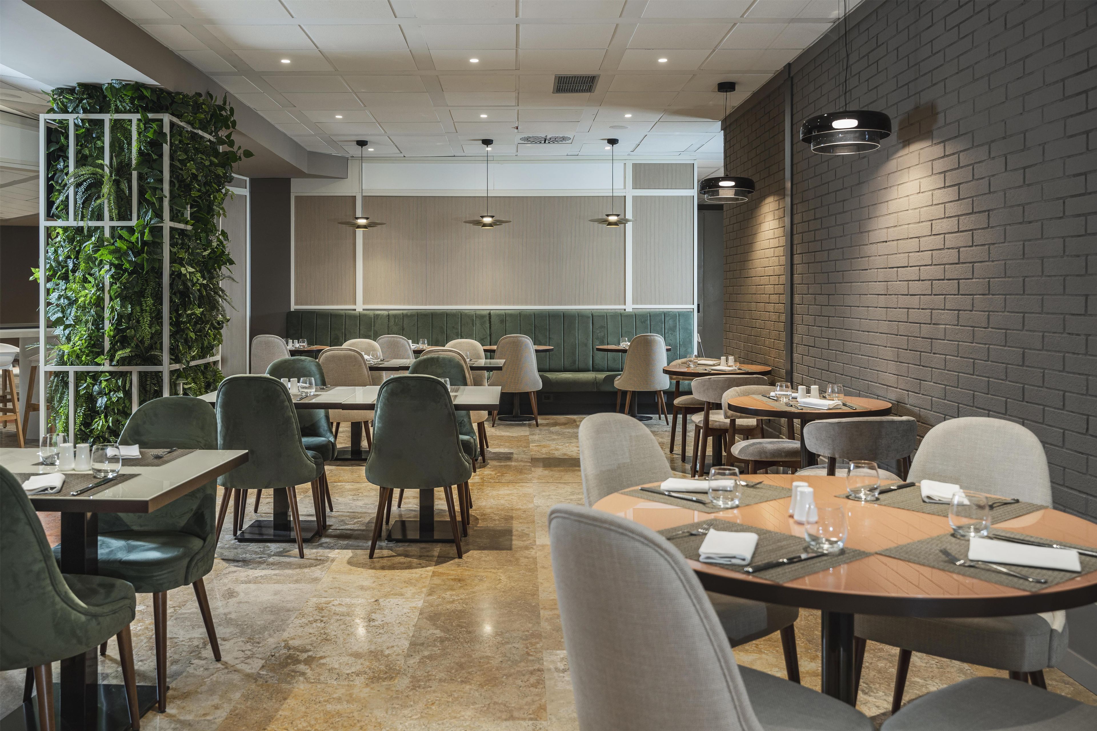 A restaurant interior with multiple dining tables and chairs, a green plant wall, and a brick accent wall.