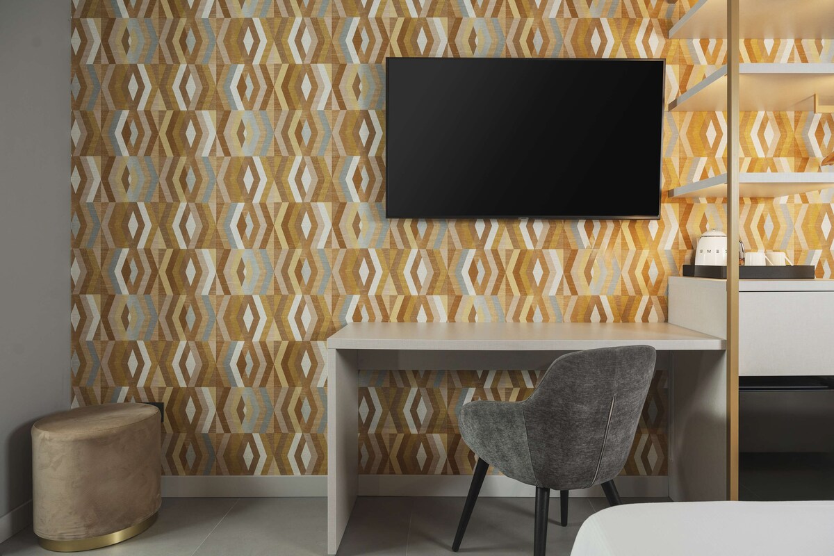Una camera moderna con carta da parati decorata, una TV a schermo piatto montata a parete, una scrivania, una sedia grigia e un comodino con una lampada.