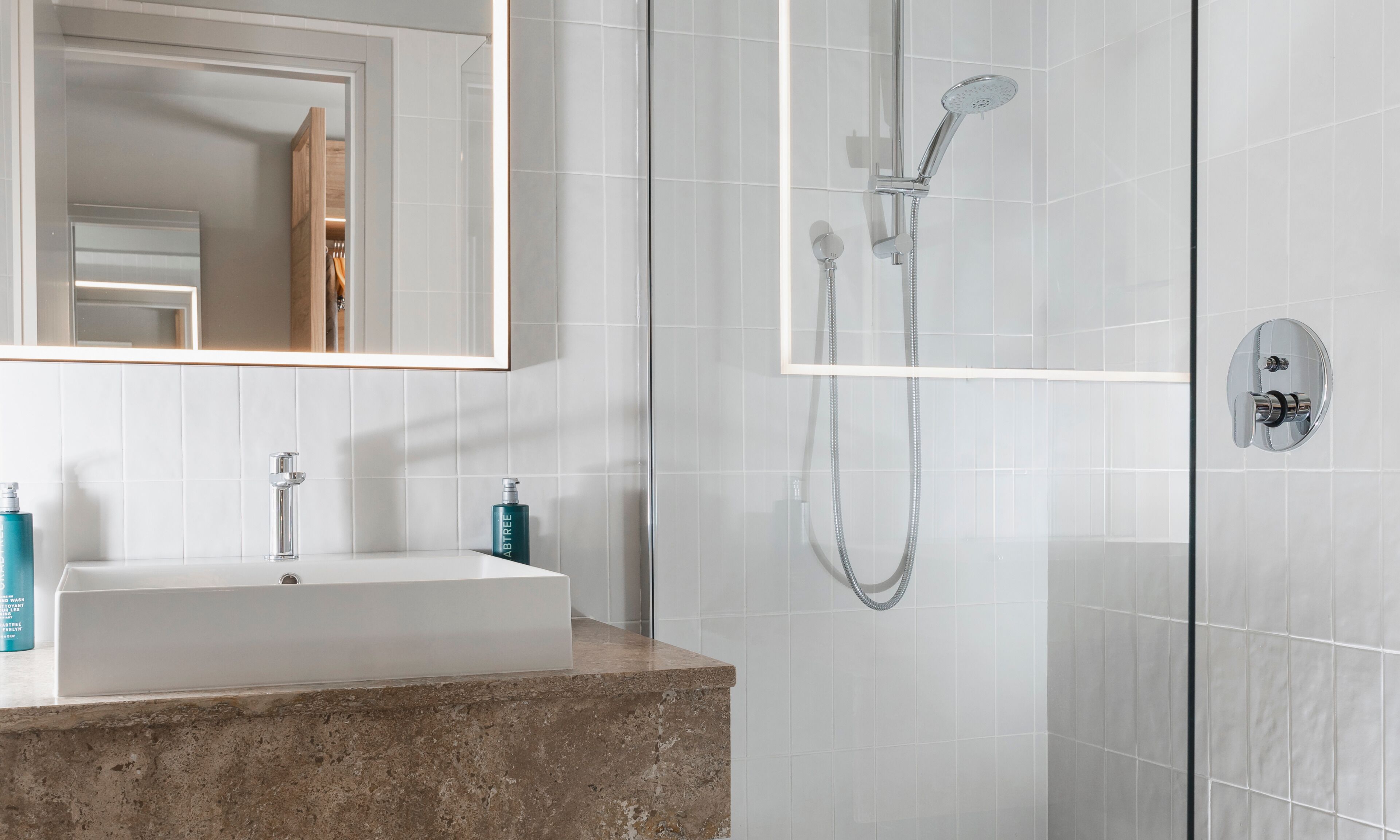 Un baño con lavamanos de mármol, un espejo, ducha con cabezal de ducha manual y una botella de jabón para manos.