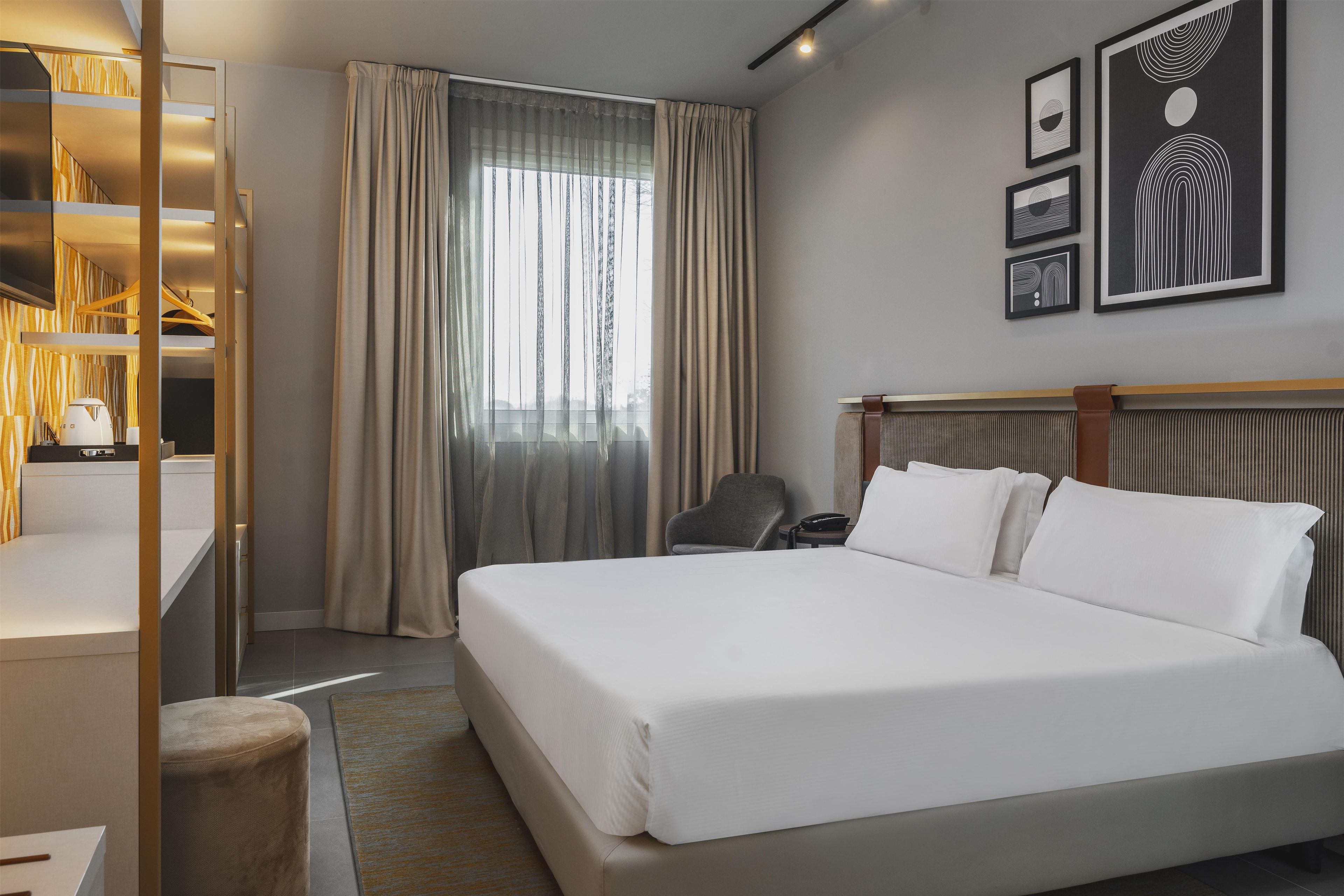 Habitación de hotel con cama, una silla, escritorio, lámpara y ventana con cortinas.