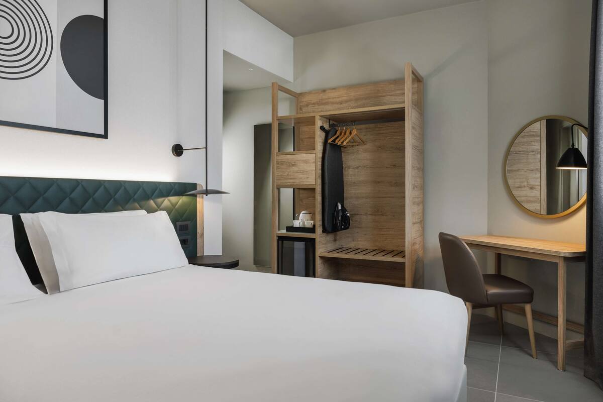 A modern hotel room with a bed, a desk, a chair, a wardrobe, and a round mirror.