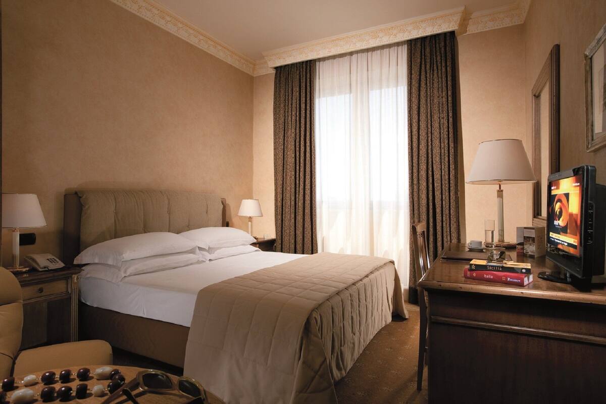 A hotel room with a bed, two bedside tables, a desk with a television, and a chair.
