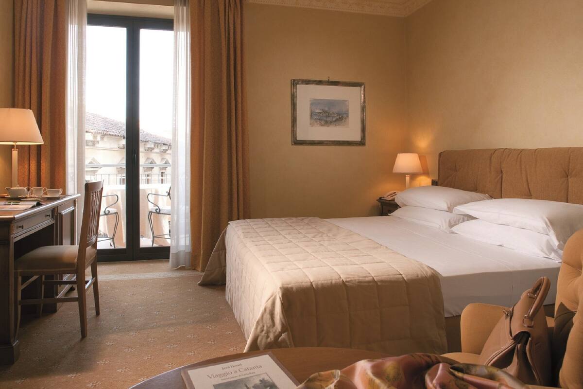 A hotel room with a bed, desk, chair, and a view of the outside.