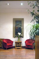 Una stanza con due poltrone rosse, un tavolino in legno, un vaso grande con fiori bianchi e un quadro appeso al muro.