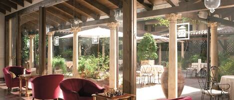 A covered outdoor seating area with red chairs, a chess table, and large windows overlooking a garden.