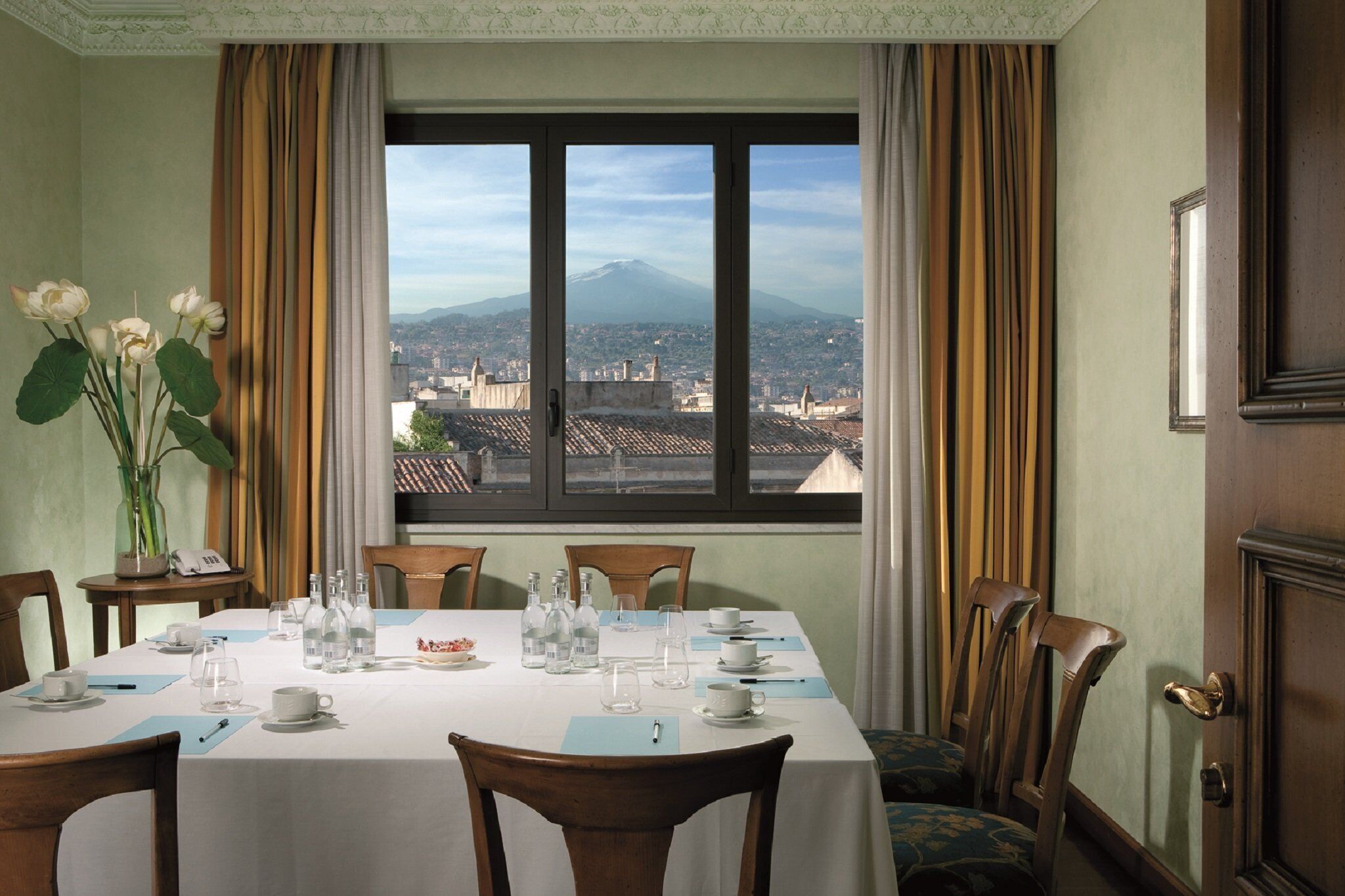 A dining room with a table set for six, a vase of flowers, and a view of a mountain through a large window.