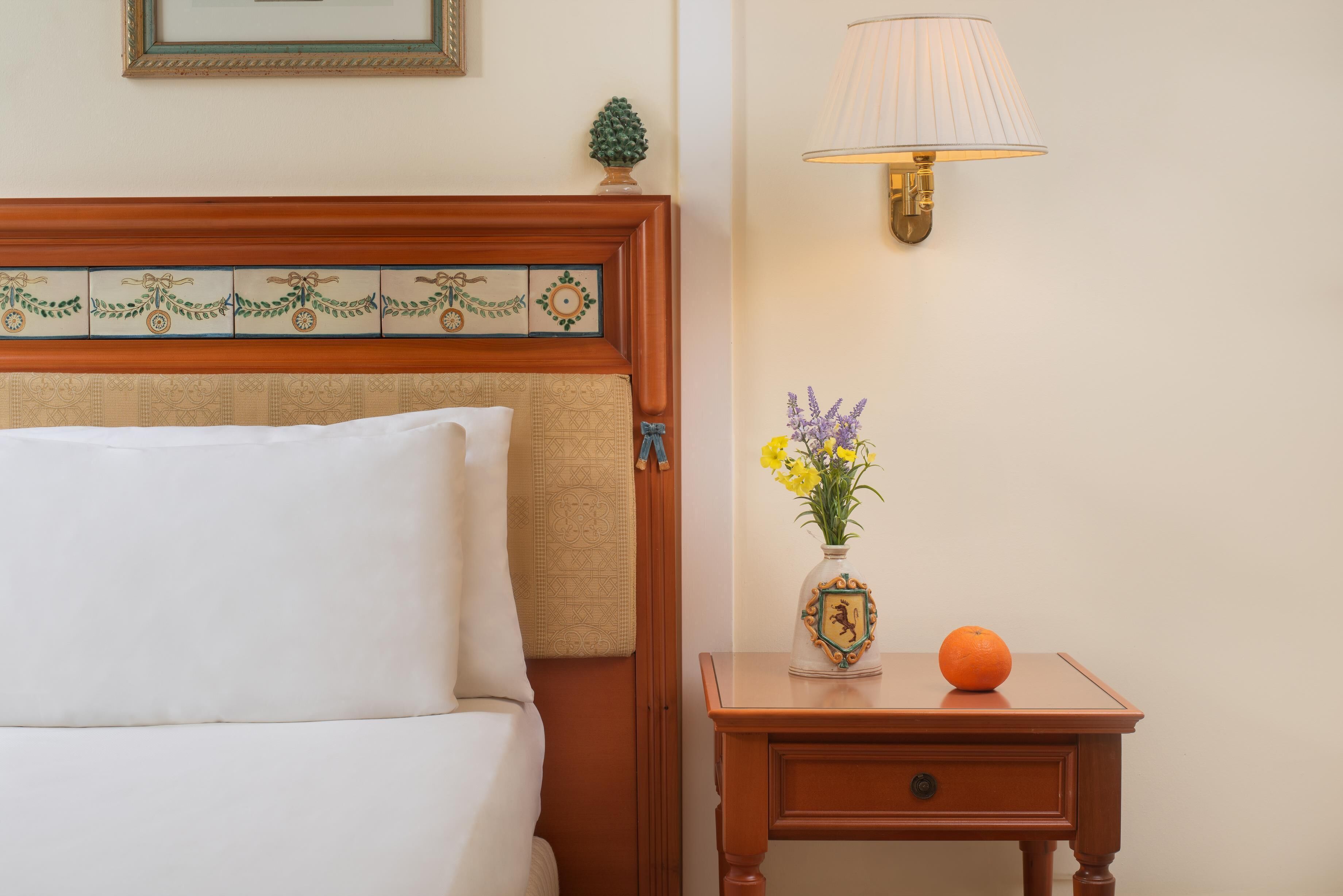 Un lit bien fait, avec une tête de lit ornée d’un motif décoratif en carreaux, une table de chevet sur laquelle se trouve un vase de fleurs et une orange, et une lampe fixée au mur.