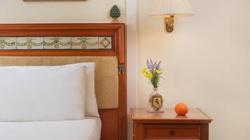 Un lit bien fait, avec une tête de lit ornée d’un motif décoratif en carreaux, une table de chevet sur laquelle se trouve un vase de fleurs et une orange, et une lampe fixée au mur.