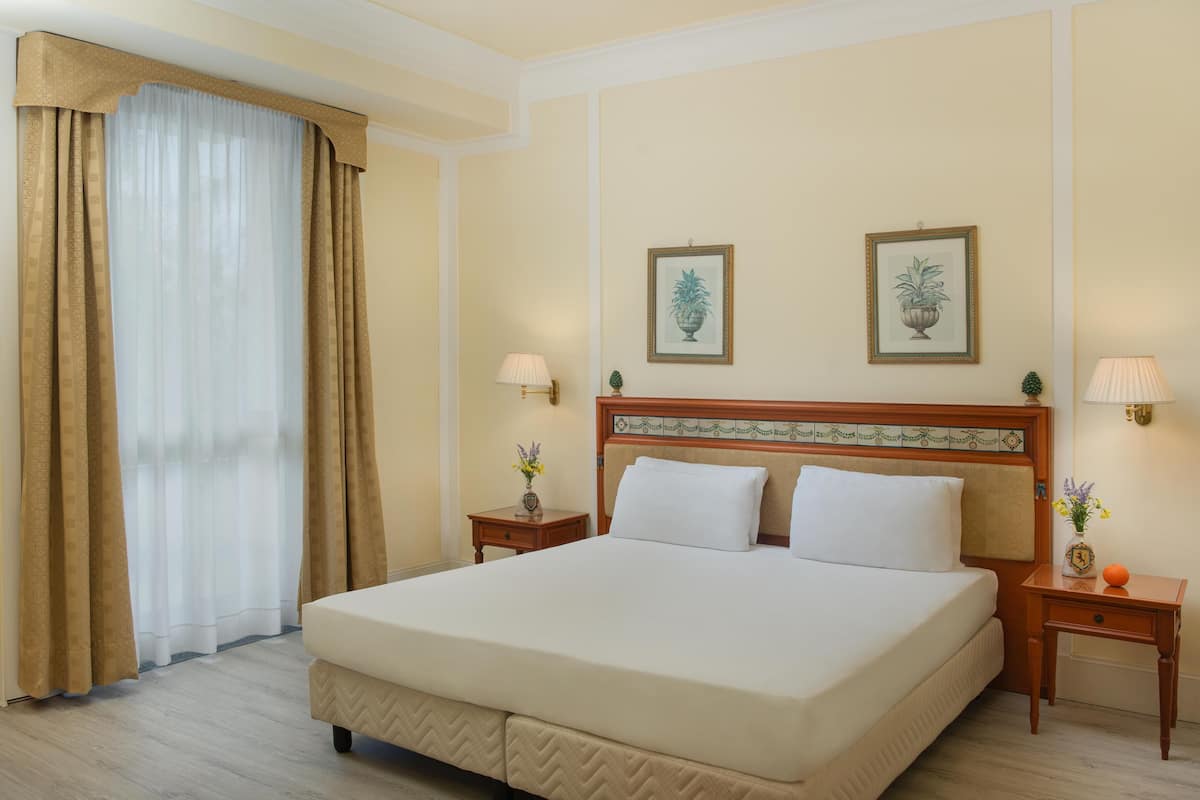 Hotelkamer met een groot bed, twee nachtkastjes met lampen, een hoofdbord met decoratief paneel en twee ingelijste foto’s aan de muur.